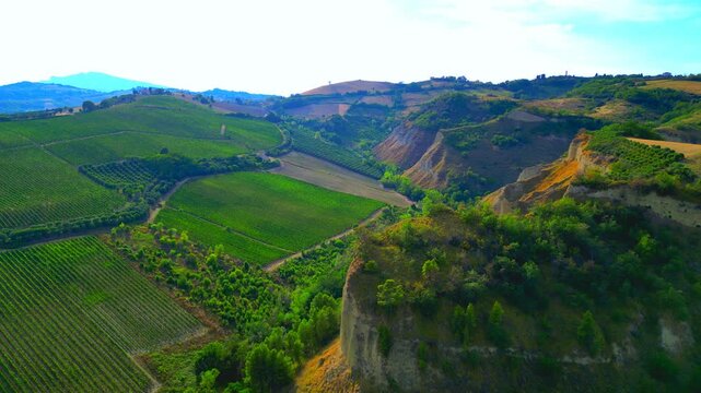 Delicate aerial push-in shot in Offida, drone flying high near Chiesa di Santa Maria della Rocca and capturing the lush surrounding valleys, striped fields, thickets, along massive green promontories