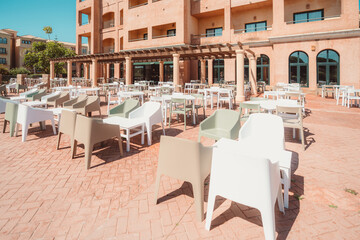 Outdoor hotel terrace with modern plastic chairs and tables arranged in rows on red brick paving, facing a Mediterranean-style building with arched windows and balconies; wide-angle shot