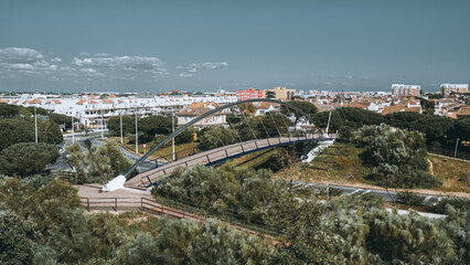 Fototapeta premium Modern pedestrian bridge with sleek metal arch crossing over green urban park, surrounded by white Mediterranean-style houses and pine trees in Punta Umbria, Andalusia, Spain