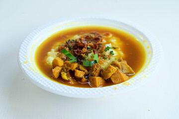 Closeup Of Savory Chicken And Rice Porridge With Yellow Sauce, Garnishes, And Herbs On A White Plate