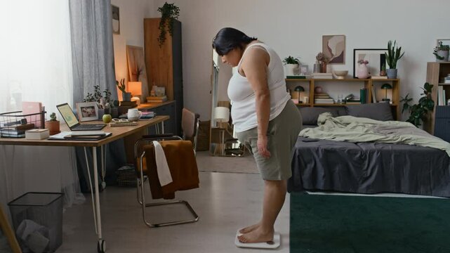 Curvy young Latin woman stepping on digital scale in minimalist bedroom after sleep