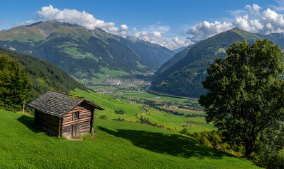 Obraz premium Idyllic valley view with cabin, rolling hills, and mountains under a blue sky with scattered clouds
