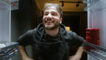 Cheerful man looking into refrigerator from inside fridge point of view at night