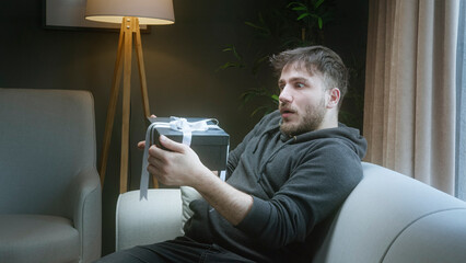 Surprised young man holding an unexpected gift box while sitting on a couch in a cozy living room