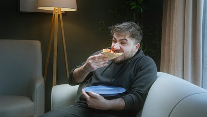 Young man relaxing on a sofa while eating pizza at home in cozy evening light