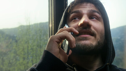 Man in hoodie talking on smartphone by window with scenic forest view on a misty day