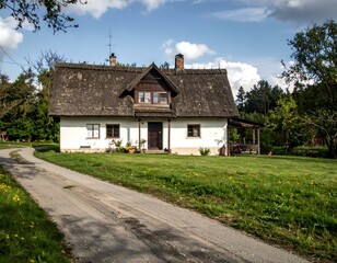 Naklejka premium Charming thatched-roof cottage on a sunny day. Countryside home with a grassy lawn and a paved road.
