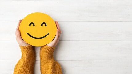 A person in a yellow sweater holds a large, smiling yellow emoji face in front of a white wooden background.