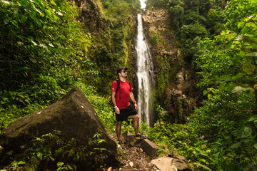Adult male hiker posing in front of a landscape with a waterfall of pure water in the middle of the mountain surrounded by tropical forest and Costa Rican jungle