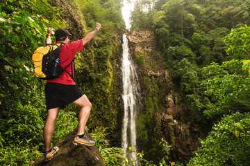 Adult male hiker posing on a rock in front of a landscape with a waterfall of pure water in the middle of the mountain surrounded by tropical forest and Costa Rican jungle