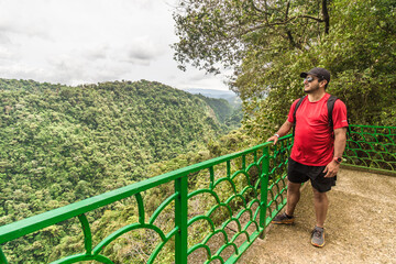 Adult man in sportswear wearing a backpack and sunglasses observing a landscape with a mountain view surrounded by tropical forest on a cloudy day in the Costa Rican jungle