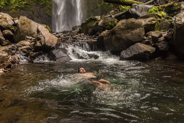 Horizontal shot of an adult man swimming in a river with a landscape in the background with a waterfall of pure water in the middle of the mountain surrounded by tropical forest and Costa Rican jungle