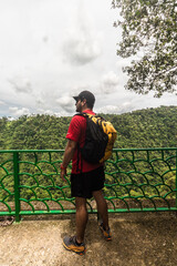 Vertical shot of an adult man wearing sportswear and a backpack looking at a landscape with a mountain view surrounded by tropical forest on a cloudy day in the Costa Rican jungle.