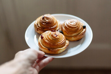 Hands holding a white plate with three freshly baked cinnamon rolls. The rolls are golden brown, swirled with cinnamon, and dotted with dried fruit, showcasing a homemade look.
