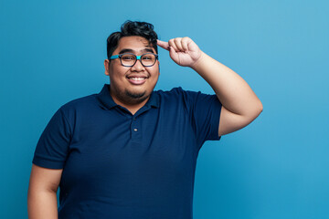 Fototapeta premium A photograph of an overweight young Indonesian man wearing a blue polo shirt and glasses, pointing at his head with one hand while smiling in the studio, isolated on a solid color background.