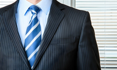 Businessman in suit with blue tie stands near a window with blinds