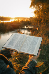 Person reading open Bible outdoors at sunset near lake, surrounded by golden light, warm tones, and...