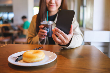 Closeup of a woman holding and using mobile phone while drinking iced coffee and eating donut at coffee shop