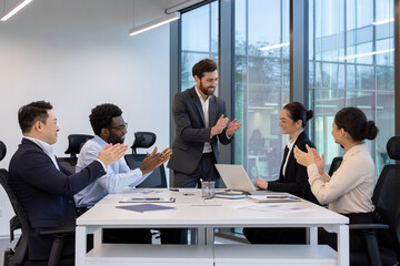 A diverse group of businesspeople clapping and smiling during a meeting in a modern office space with natural light.