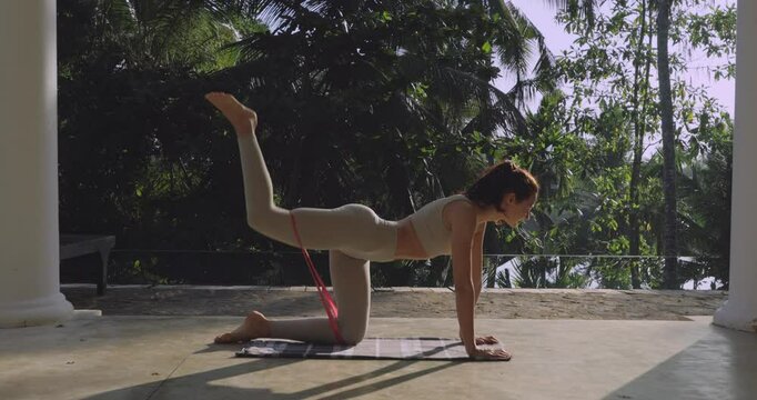 A fit woman works out with a latex resistance band, targeting her glutes and legs in a tropical villa. Sunlight streams through palm trees as she embraces fitness and body toning. 