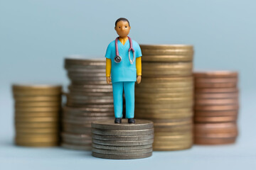 A small doctor figurine stands on stacked coins, with larger coin stacks blurred in the background, symbolizing healthcare costs and financial concerns in medicine.
