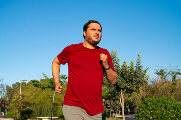 A man in a red shirt jogs through a park filled with trees and shrubs under a bright blue sky. His...