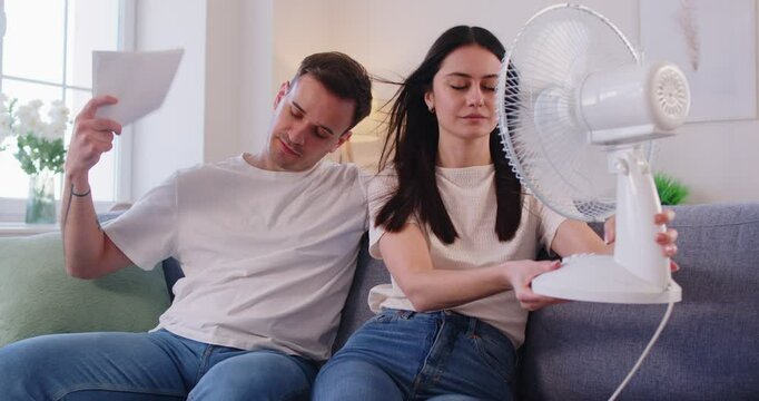 Family couple posing in room with electric fan, suffering from heat during summer weather, being in overheated apartment, sitting on sofa in front of stream of cold air cooling themselves