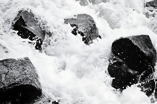 A high contrast black and white image of a rugged beach with large rocks and foamy waves crashing against them