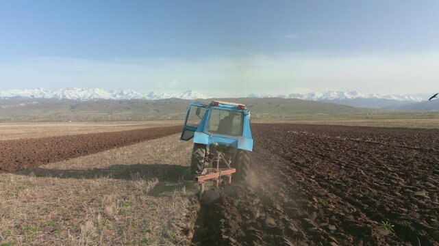 Blue Tractors sowing on agricultural field in spring, readying the field for planting. Powerful tractor turns the soil against the backdrop of high mountains. Issyk-kul Region valley in Kyrgyzstan