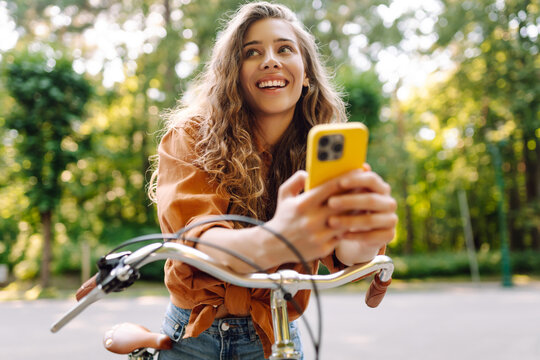 Smiling woman with phone riding bicycle on sunny street in park. Young woman enjoying sunny day and having fun outdoors. Weekend and blogging concept.