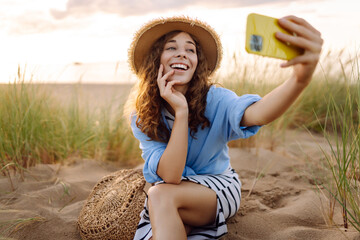 Cheerful woman in hat and modern clothes and phone enjoys sunset on beach. Beautiful tourist takes...