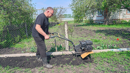 A man is using a rototiller in a yard. The man is wearing a black shirt and black pants