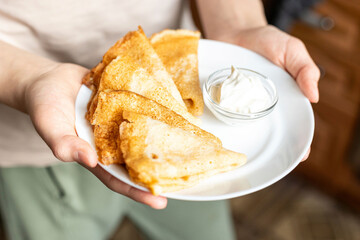 Plate of golden, folded crepes served with a small bowl of sour cream. A hand is seen gently...