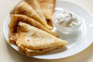 Plate of golden, folded crepes served with a small bowl of sour cream. A hand is seen gently holding the plate, showcasing the freshly made snack.