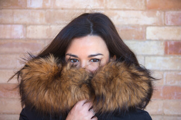 Young and beautiful brunette woman with black coat and fur hood covers her neck and holds it with...