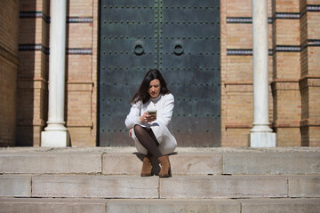 Young and beautiful Latin brunette woman is sitting on a staircase looking at her mobile phone. The...
