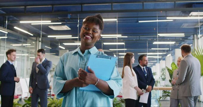 Portrait of happy smiling african american business woman leader proudly looking at camera holding documents in hands. Female company employee standing in modern office. 4k video. slow motion video.
