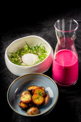A stylish presentation of a gourmet meal featuring a bowl of fresh salad with herbs and a soft egg, accompanied by roasted potatoes and a vibrant pink drink in a glass pitcher.