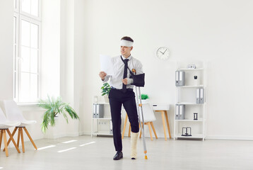 Man with a broken arm and leg in bandage stands with crutches, working in the office with...