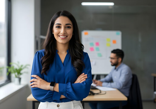 confident young businesswoman in blue shirt standing with arms crossed in modern office environment with colleague in background
