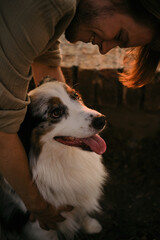 Tender moment between a young man and his Australian Shepherd dog at sunset. The man leans in smiling, while the dog looks up with trust and affection, bathed in warm golden light.