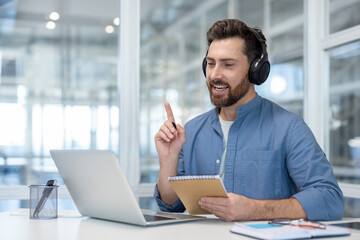 Obraz premium A man in a blue shirt with headphones gestures while on a video call, holding a notepad, indoors at a bright office.