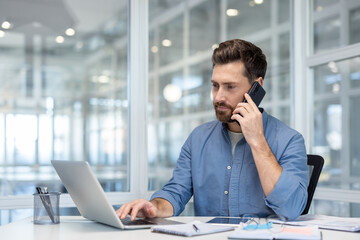 A focused businessman works on a laptop while talking on his smartphone in a modern office setting, multitasking.