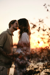 Romantic young couple standing close and smiling at sunset, holding hands behind soft-focus branches. The dreamy composition and golden light evoke warmth, intimacy, and connection.