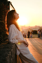 Side profile of a young woman leaning against a stone wall at sunset, looking thoughtfully toward...