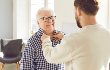 Adult son helping his senior father to put on his clothes. Young man buttons up the shirt of his...