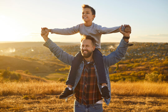 Happy smiling child boy sitting on fathers shoulders holding hands and looking into the distance enjoying sunset and beautiful nature in the field. Love,care and Fathers day concept