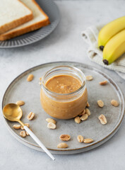 Peanut butter in glass jar on light  background with salted nuts, banana and bread toasts.