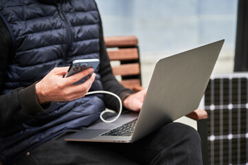 Cropped view of man sits on wooden bench, working on laptop and smartphone, both connected to solar...