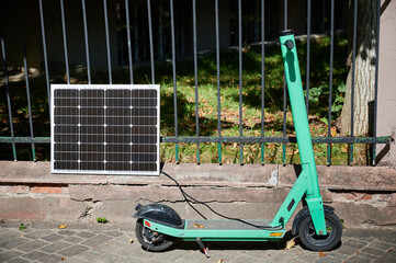Mint-green electric scooter parked on stone pavement next to solar panel leaning against iron...